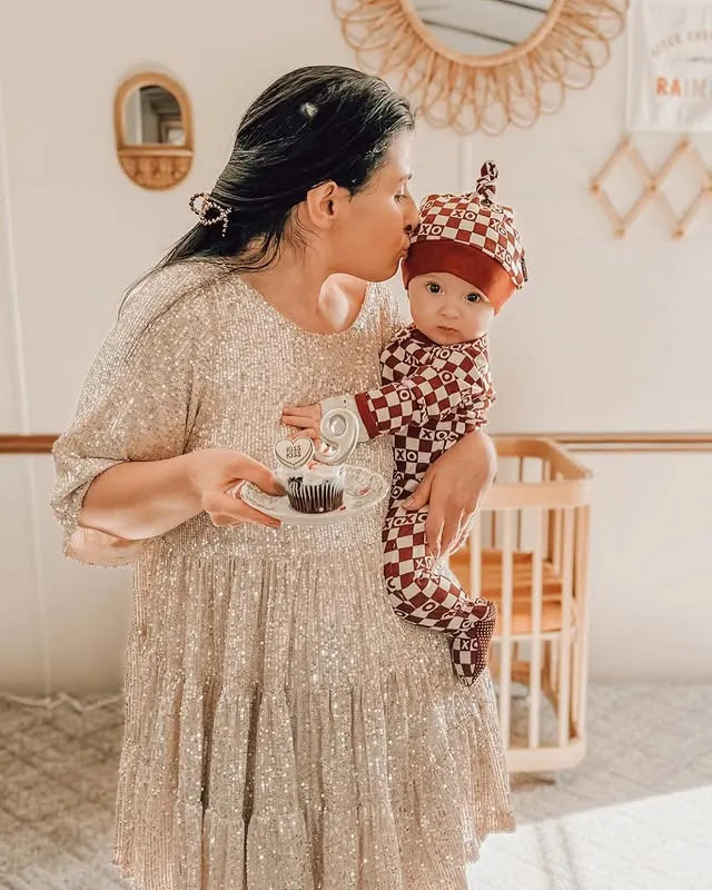 Woman in a sparkly dress holding a baby in a checkered outfit in a home setting.
