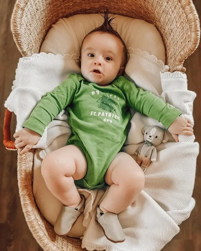 Baby in a green St. Patrick's Day onesie lying in a crib with a teddy bear.