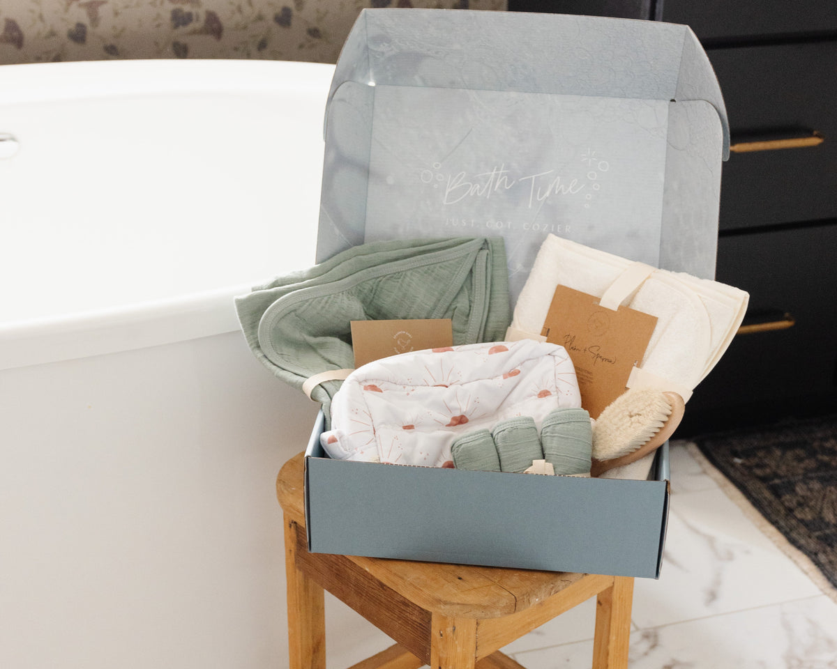 A curated baby gift set in a dusty blue box, displayed on a wooden stool next to a white bathtub. The set includes a sage green cotton bath towel, a bamboo bath towel, a sun organizer bag, wash cloths, and a small wooden brush.