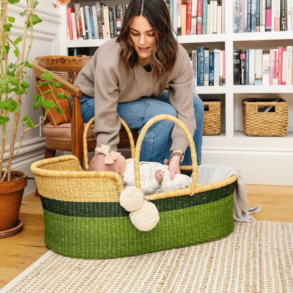 Woman holding a baby in a green and beige Moses basket in a home setting with bookshelves in the background.