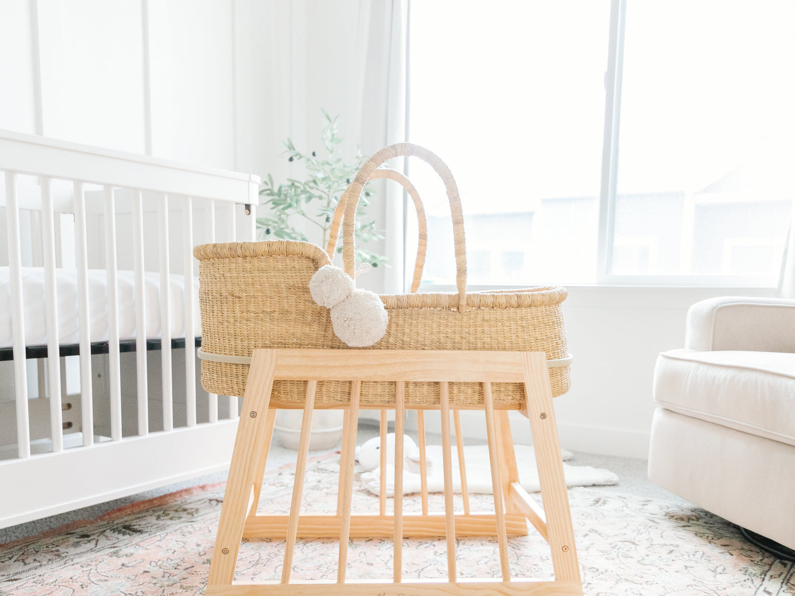 A natural woven Moses basket with two cream pom-poms sits on a light natural wood bassinet stand in a bright nursery setting.