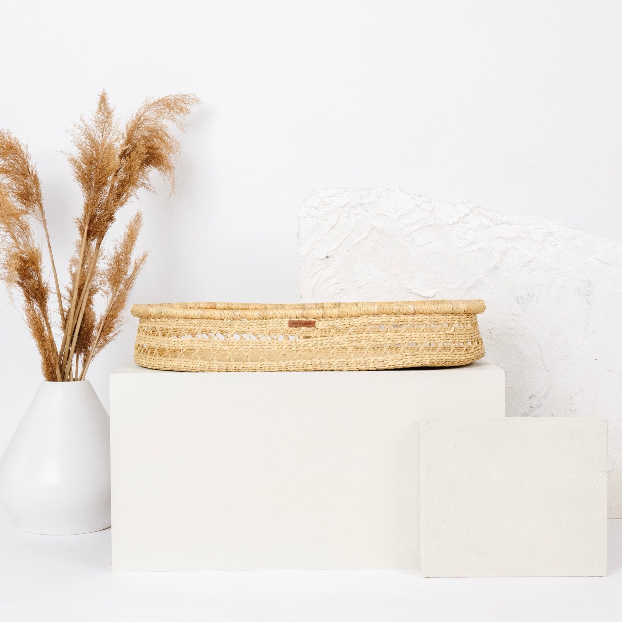 Woven basket on a white surface with a white vase containing dried pampas grass.